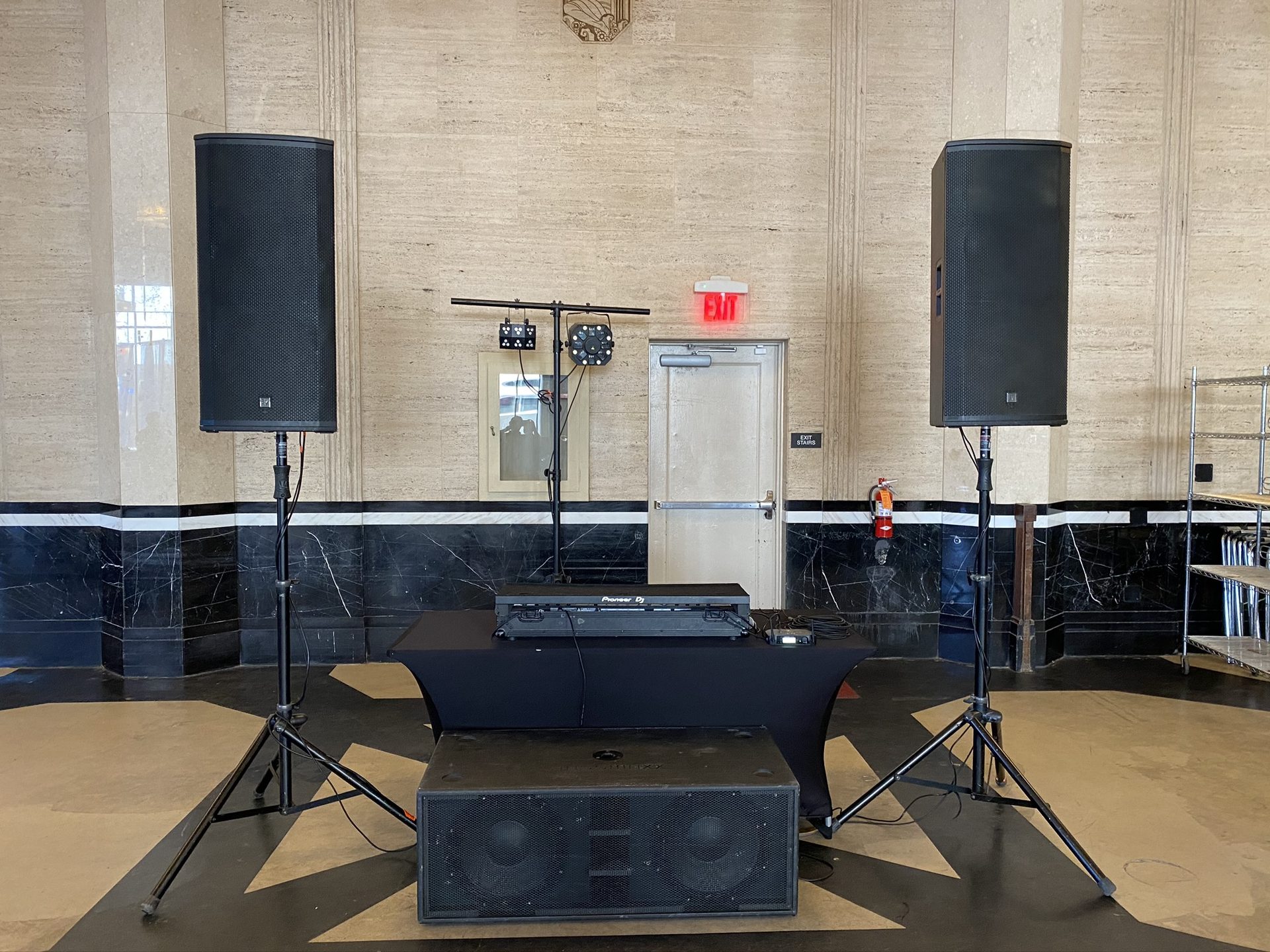 Two BassBoss DV12 speakers on stands over a subwoofer under an empty DJ booth in a Dallas venue